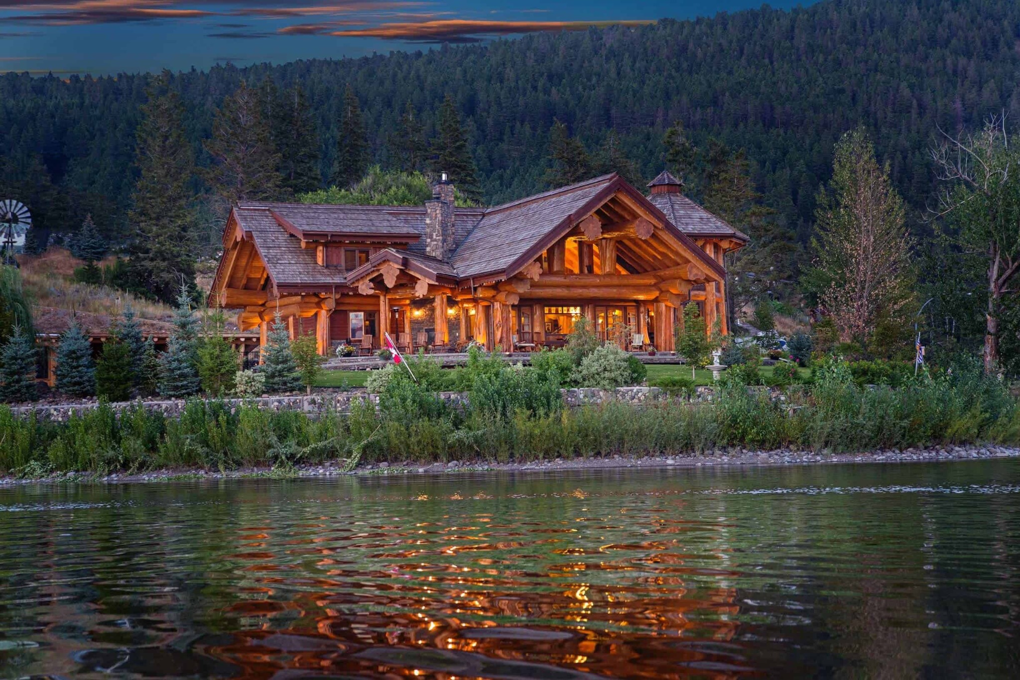 A large log home glowing at dusk beside a forested lake, with its lights reflected in the water